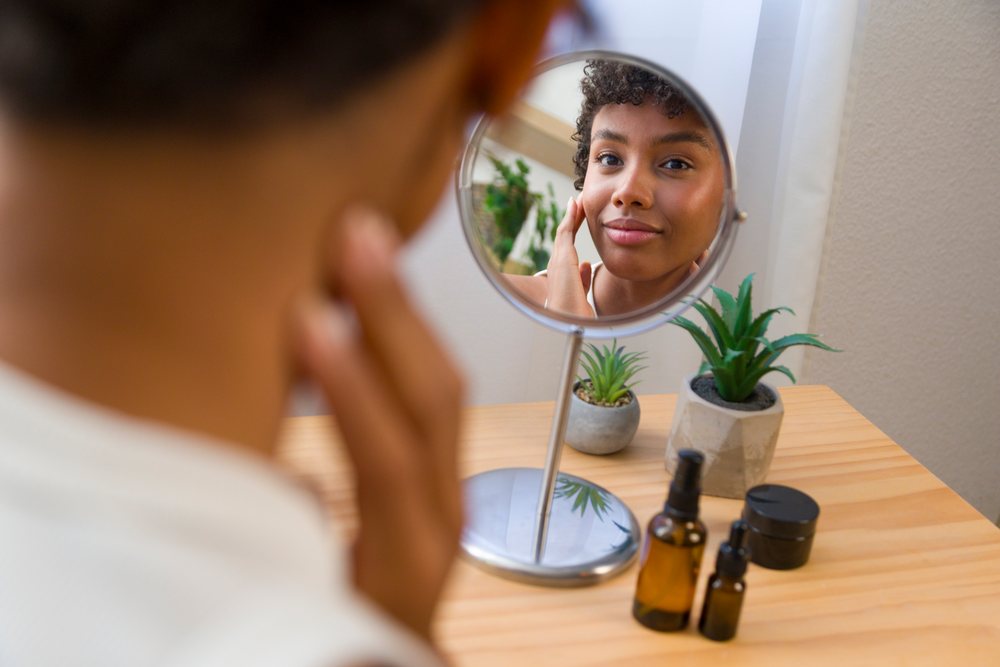 Young woman with curly hair applies facial moisturizer while looking into a mirror on a wooden table with small plants.