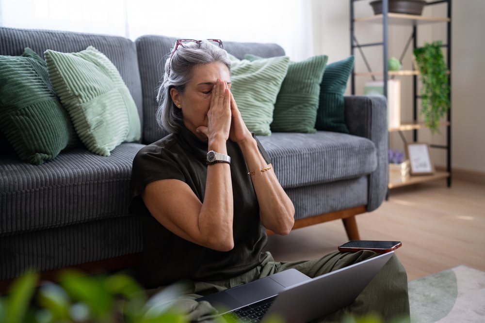 Woman with gray hair and glasses sits on the floor by a gray sofa with green pillows, using a laptop and smartphone in a home setting, Spain.