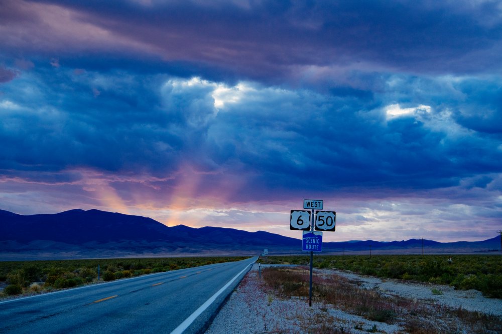 Westbound highway 6 and 50 stretch toward distant mountains under a colorful sunset sky in Nevada desert, USA.