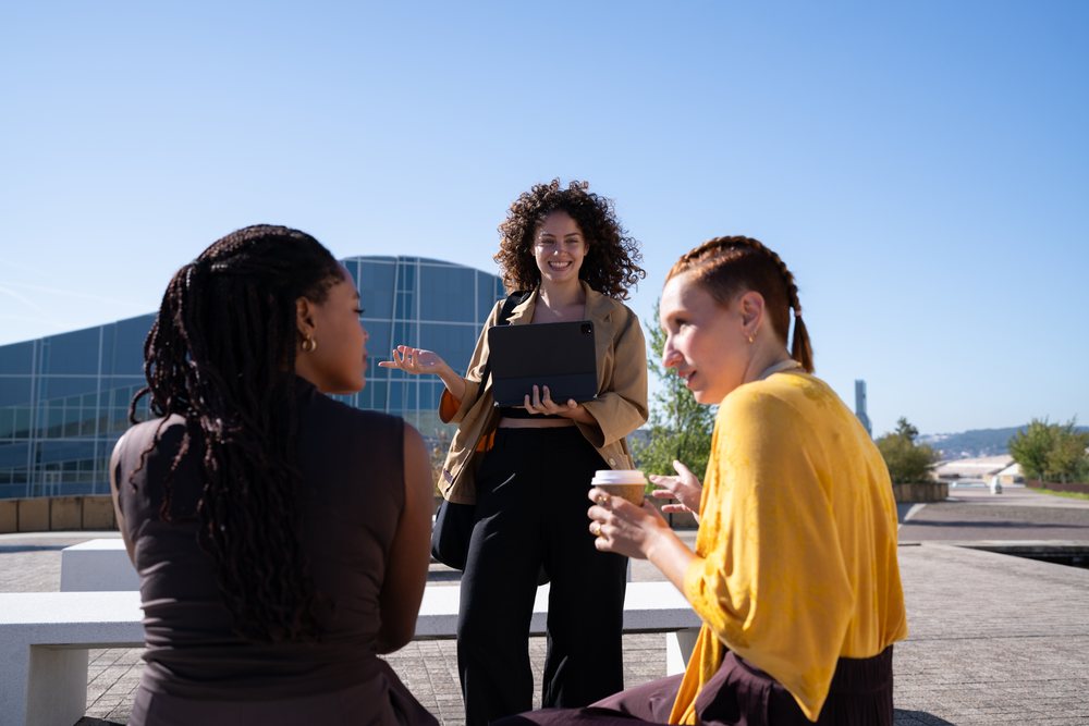 Three adult women converse outdoors near a modern glass building, engaging with a tablet and coffee on a sunny day, Spain.