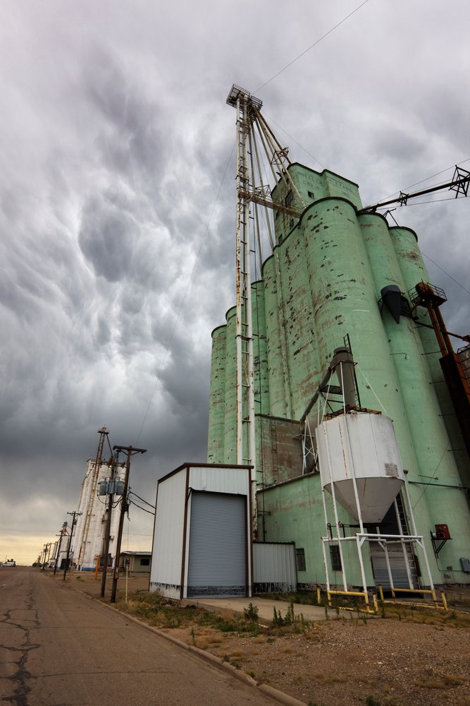 Large green grain elevator with multiple storage silos stands under stormy clouds along a rural cracked road, Texas, USA.
