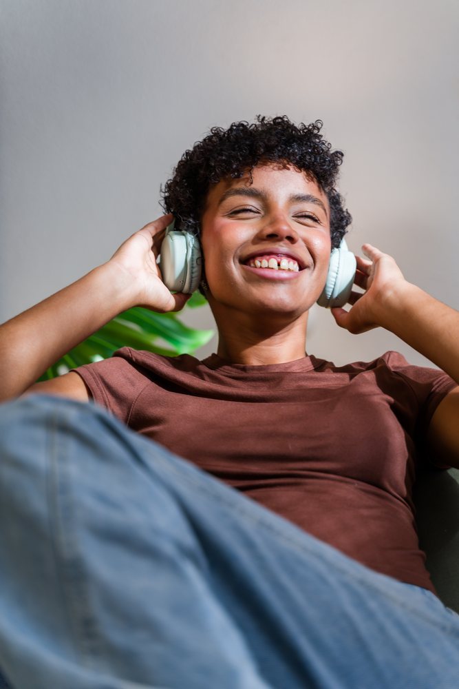 Smiling woman with curly hair listens to music with over-ear headphones indoors beside a green plant leaf, Spain.