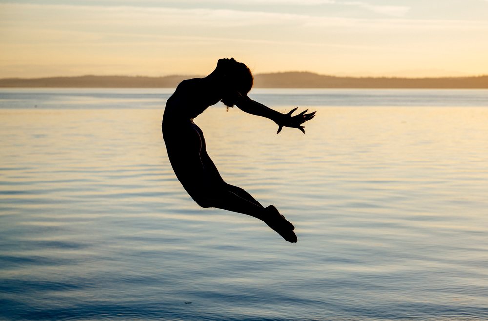 Woman performing an acrobatic jump in silhouette against a sunset over a calm lake, yoga, acrobatics, USA.
