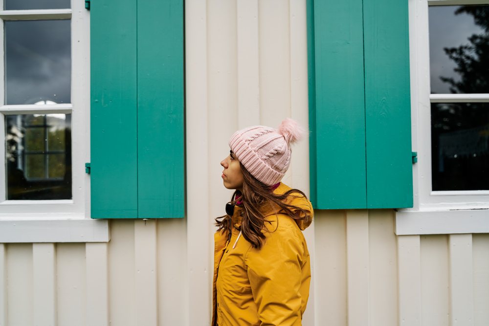 A woman wearing a yellow jacket and pink knit hat stands beside a building with green shutters and white siding, Iceland.