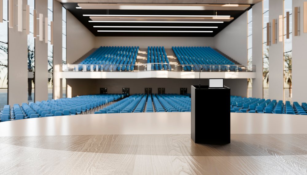 Modern auditorium's conference room features a podium with microphone and laptop facing rows of blue chairs and balcony seating during daytime.