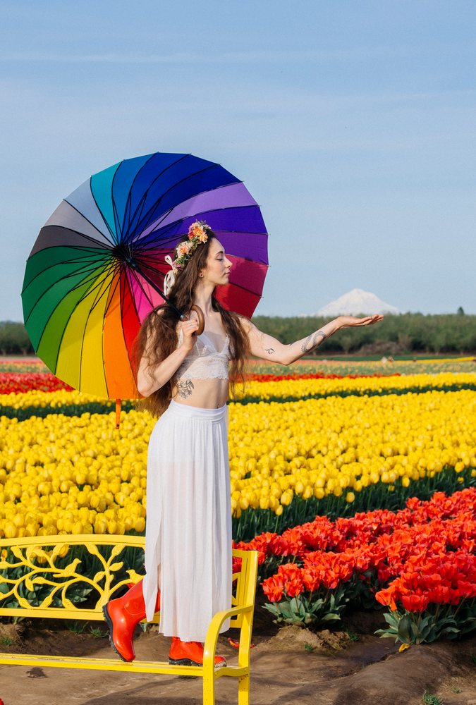 Woman standing on a yellow bench in a tulip field holding a rainbow umbrella with a flower crown and mountain view, USA.