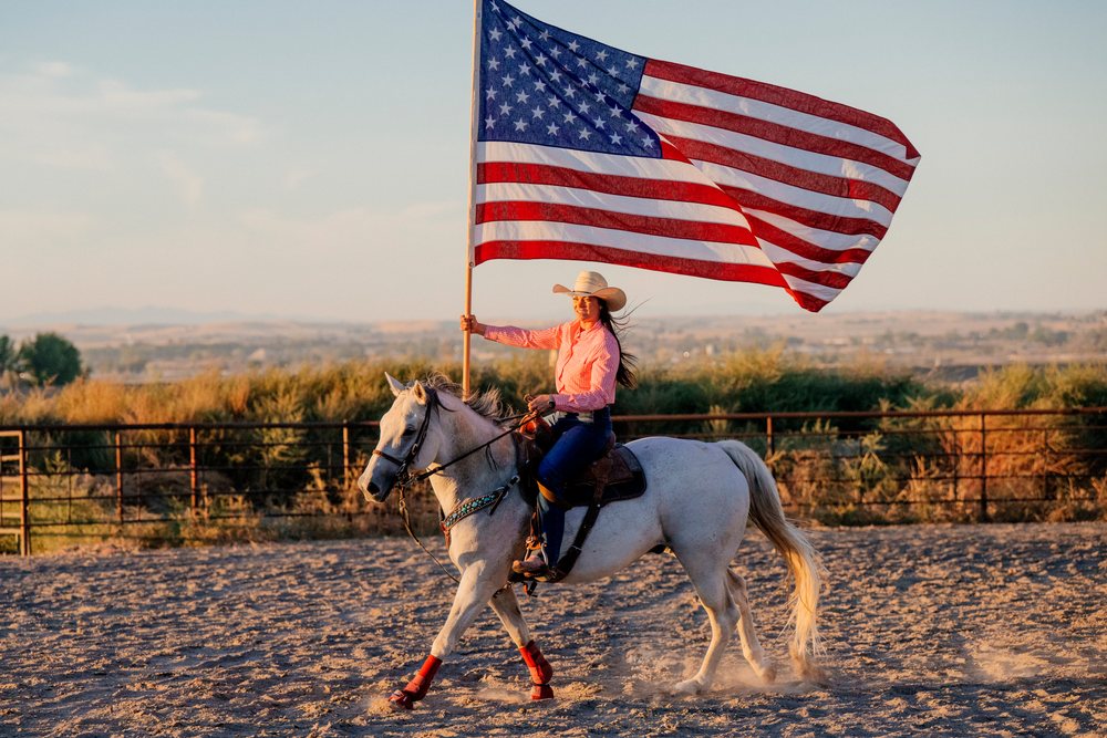 Woman riding a white horse in a sand riding arena holding an American flag at sunset, Southwest USA.