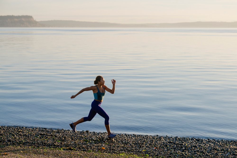 Woman running along a rocky beach shoreline with calm water, yoga, acrobatics, USA.