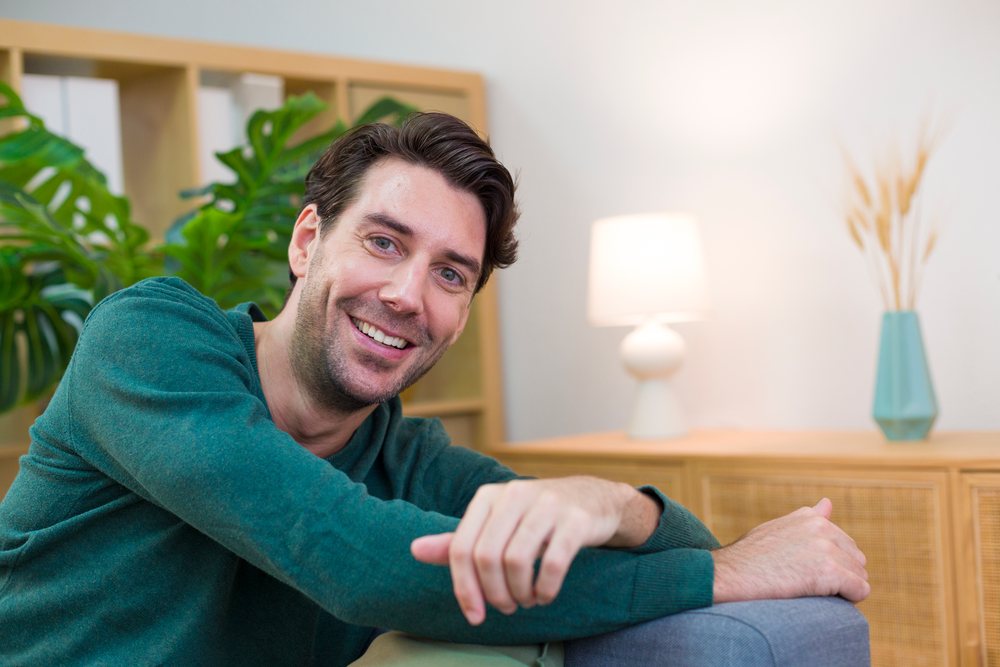 Man wearing a green sweater smiles while relaxing in a cozy indoor living room with a plant, lamp, and bookshelf, Technology, young adult, home.