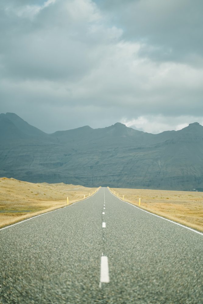A straight asphalt road with yellow roadside markers stretches through dry grassy fields toward a mountain range under a cloudy sky, Iceland.