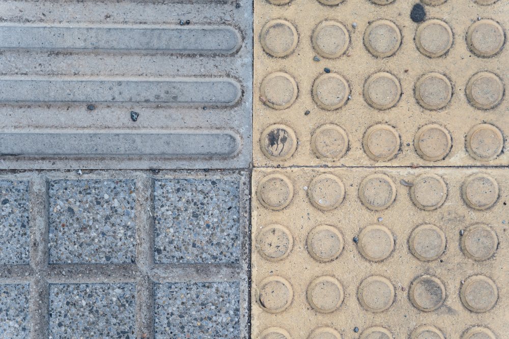 Textured yellow tactile paving with circular dots and gray grooved concrete tiles side by side on an outdoor urban sidewalk, texture, abstract texture, background, wallpaper, Spain.