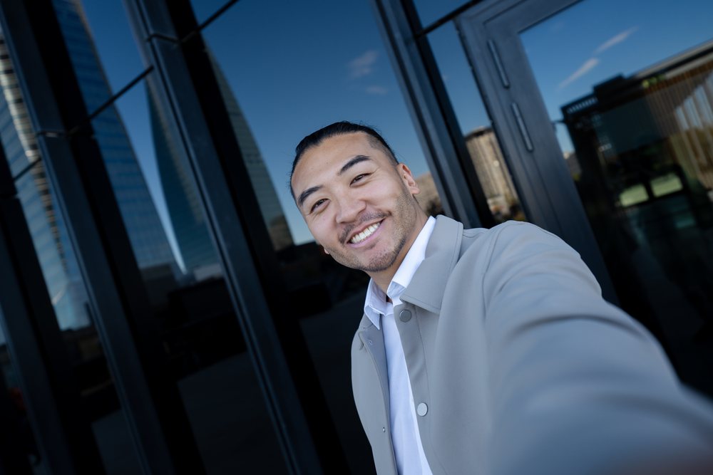 Man wearing a gray jacket smiles while taking a selfie outdoors in an urban area with reflective glass buildings, Madrid, Spain.