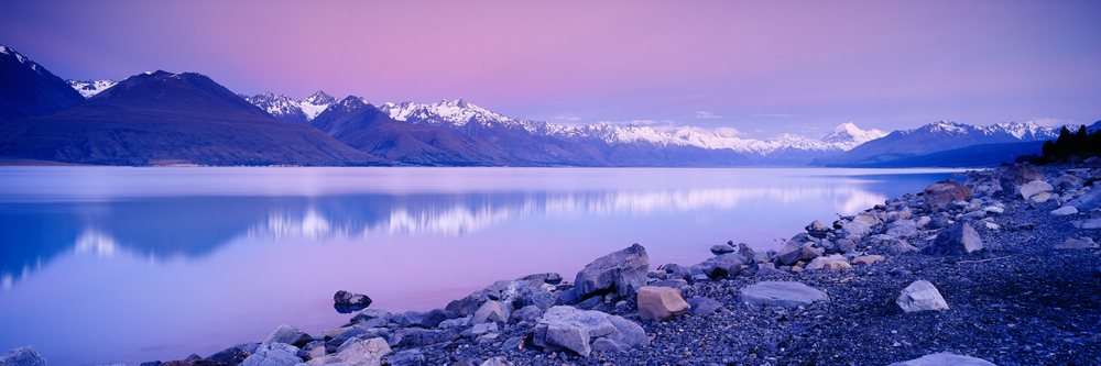Mountains with snow reflected in the calm lake by a rocky shore at dusk.