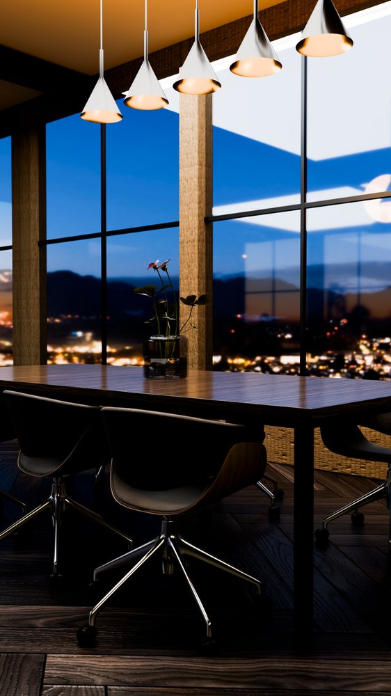 Modern wooden dining table with black office chairs and pendant lights overlooks a nighttime cityscape through floor-to-ceiling windows.