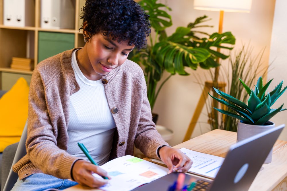 Woman with curly hair writing in planner at a wooden desk with laptop and indoor plants in a home office setting, Spain.
