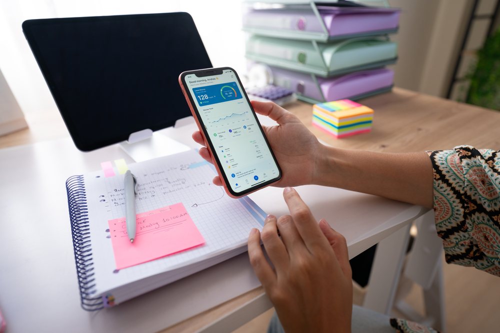 Person holding a mobile phone displaying a glucose monitoring app at a desk with a spiral notebook, pen, sticky notes, and desktop tablet in a home office, Spain.