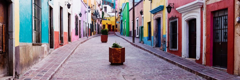 Colorful buildings line a narrow cobblestone street with potted plants and street lamps, Guanajuato, Mexico.