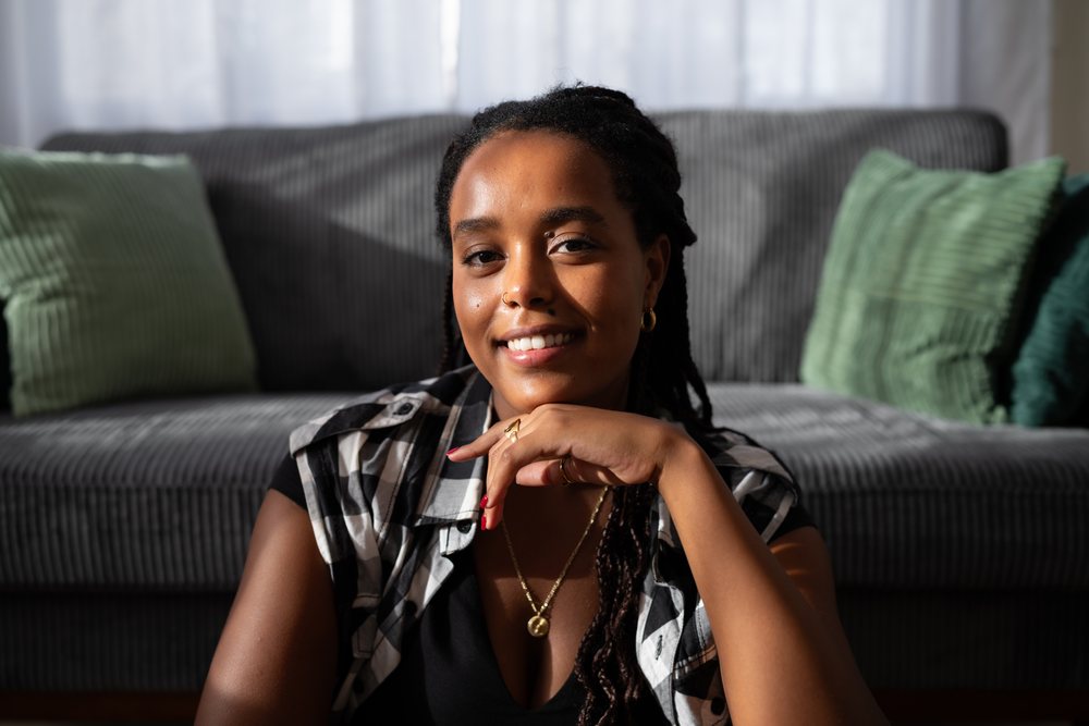 A young woman with braided hair smiles warmly while sitting indoors on a gray couch with green pillows, moving, Spain.