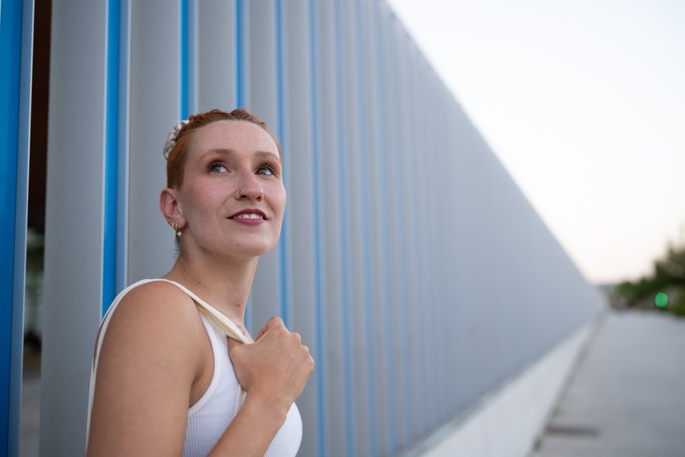 A young woman with red hair looks up thoughtfully while standing beside a blue and gray modern vertical wall in an urban outdoor setting, Santiago De Compostela, Spain.