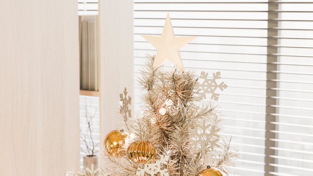 Indoor Christmas tree decorated with a star, gold baubles, and snowflake ornaments by window blinds.