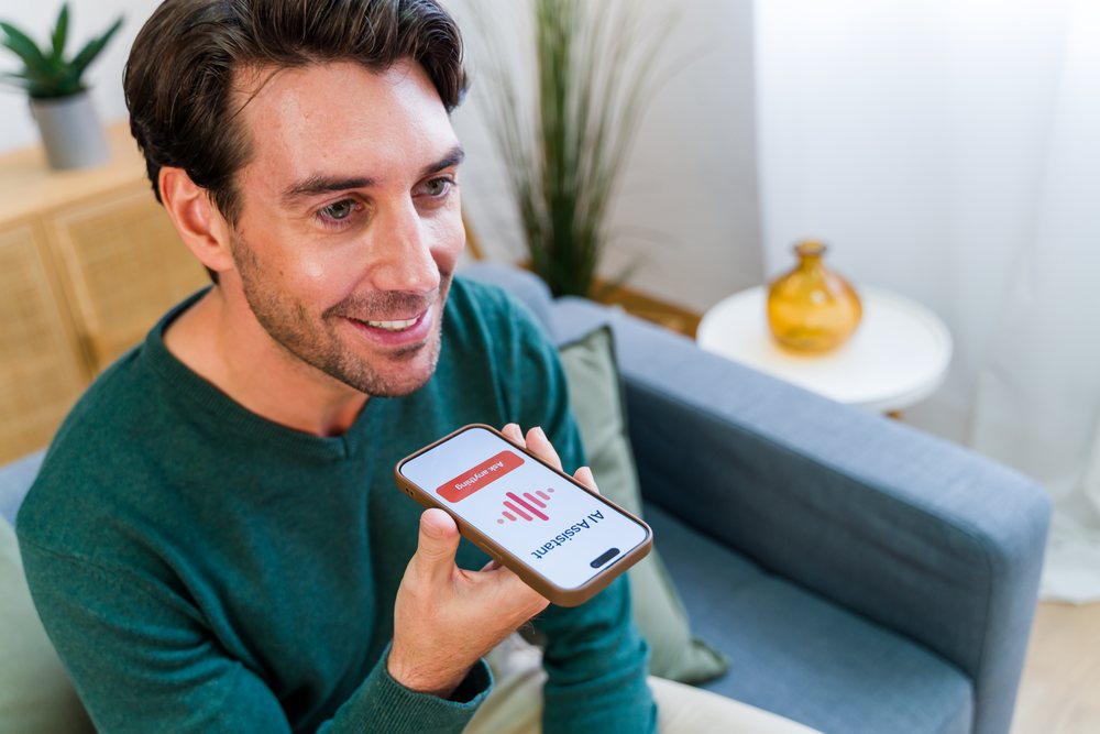 Man sitting on a couch in a living room using smartphone voice command with AI assistant, Spain.