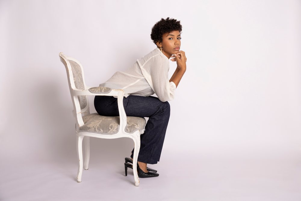 Woman with short curly hair sits thoughtfully on a vintage chair in a minimalist studio fashion portrait, young culture.