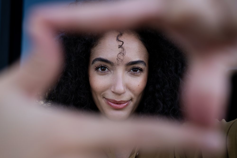 Close-up portrait of a smiling woman with curly hair framing her face with hands outdoors in natural light, Madrid, Spain.