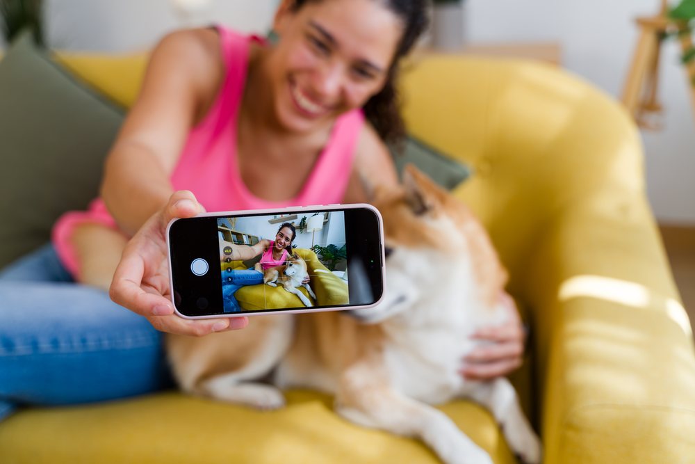 A woman happily takes a selfie with her dog on a yellow couch in a living room, Spain.