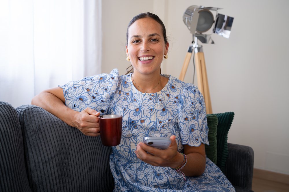 Woman in a blue floral dress smiles while holding a mug and smartphone, sitting on a dark gray sofa indoors with a lamp behind her, telecommuting, Spain.