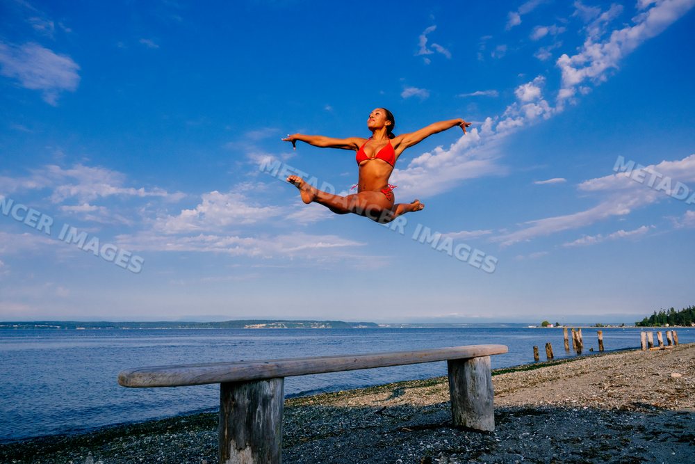 A woman jumps in a red swimsuit on a pebble beach under blue sky