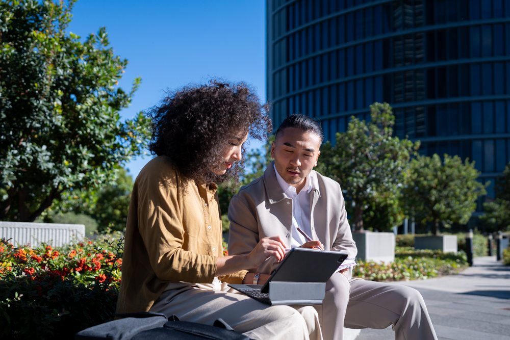 Two people collaborating outdoors with a tablet and writing pad near urban greenery and a modern office building, Madrid, Spain.