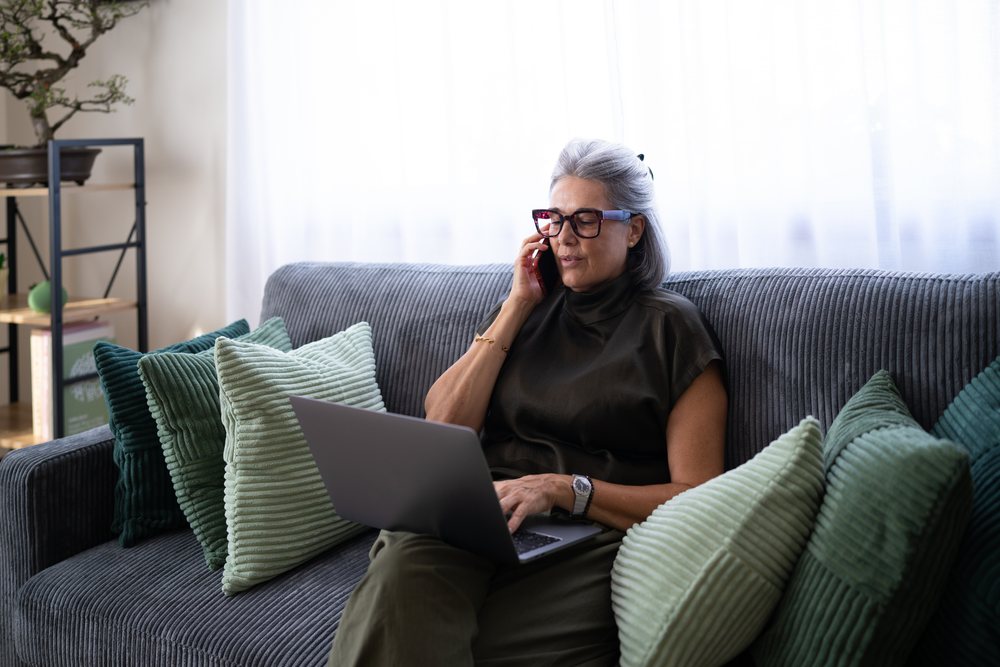 Woman with gray hair sits on a couch indoors using a laptop and talking on the phone, Spain.