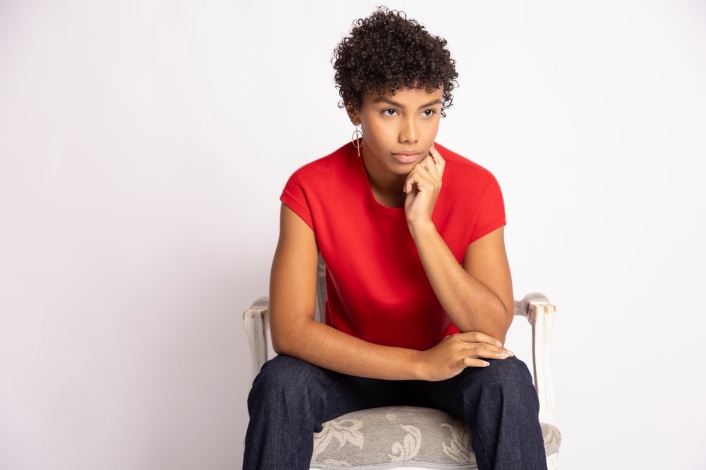 Young woman with short curly hair wearing a red shirt sits thoughtfully on a patterned chair in a studio setting, fashion, clothes, young culture.