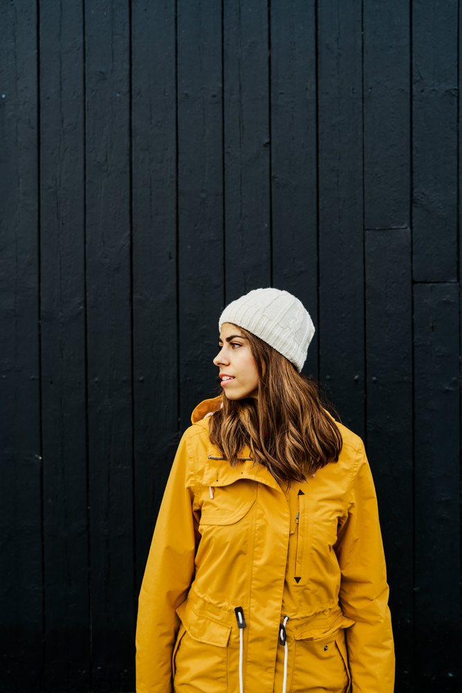 A young woman wearing a yellow jacket and white knit hat stands against a black wooden wall, Iceland.