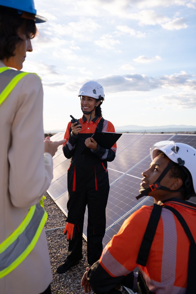 Workers wearing helmets and safety vests inspect solar panels outdoors under a clear blue sky, Madrid, Spain.