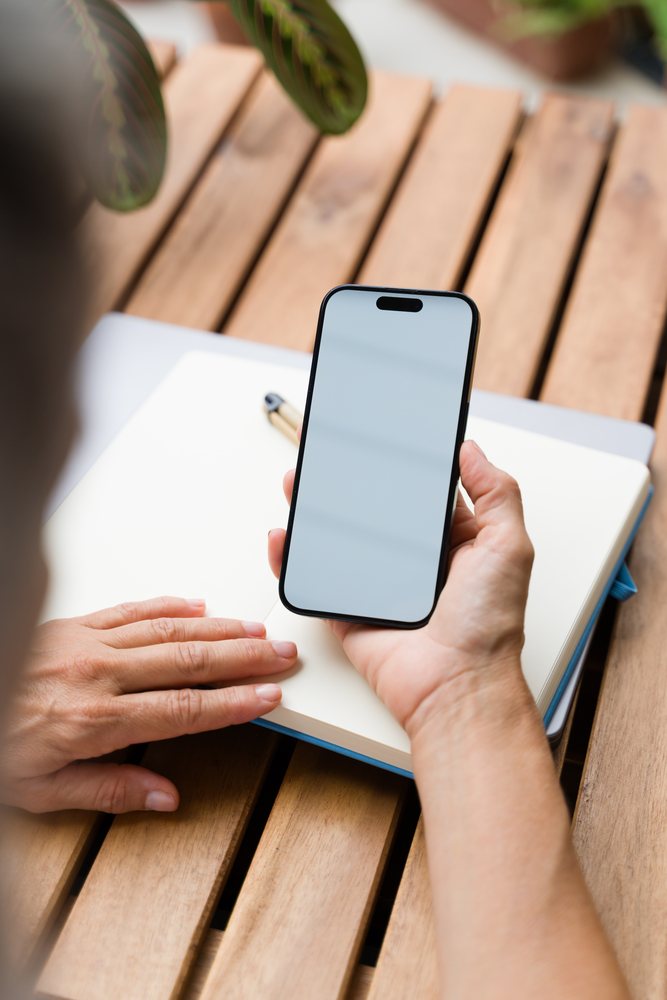 Person holding a smartphone with blank screen over an open notebook on a wooden table, Spain.