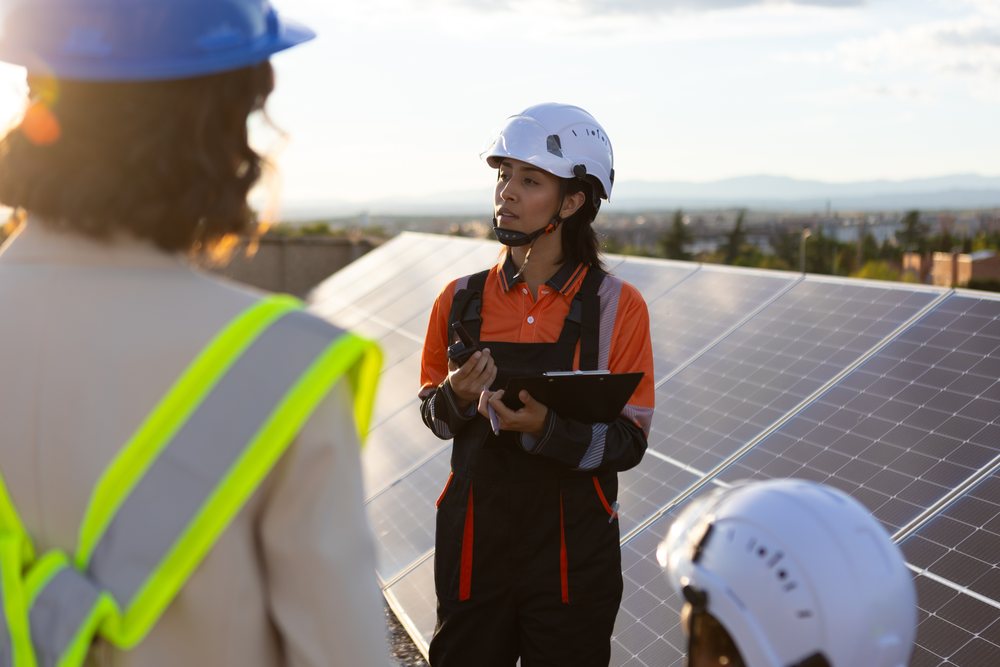 Female engineer in safety helmet and orange work uniform holding clipboard and radio device while inspecting outdoor solar panels at sunset in urban, Madrid, Spain.