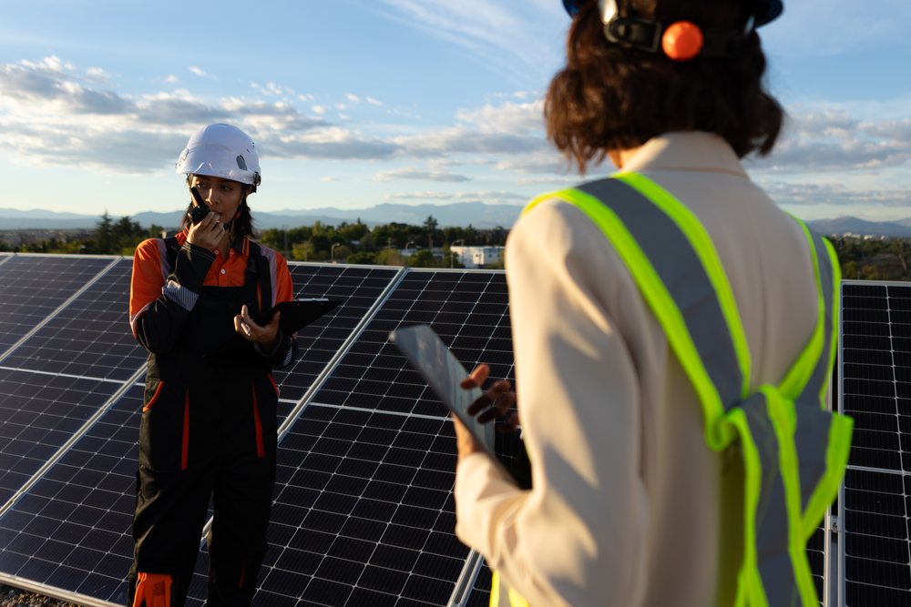 Two workers inspecting solar panels and communicating outdoors during daylight with mountains visible, Madrid, Spain.