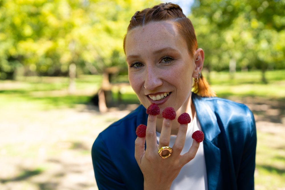 Woman outdoors in a park smiles with fresh raspberries placed on her fingers in daylight, Spain.