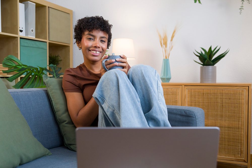 Smiling woman wearing blue jeans and a brown shirt sits on a sofa holding a mug in a living room with potted plants and green cushions, Young woman, home.