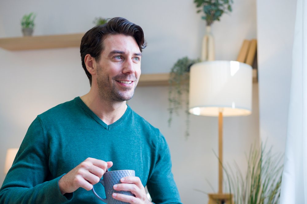 Man sitting indoors at home holding a ceramic mug and smiling, next to a lamp and potted plants, Technology, young adult.