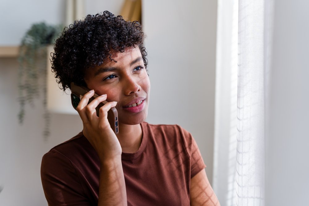 Woman talking on the phone indoors by a window with curtain and nearby plant, Spain.