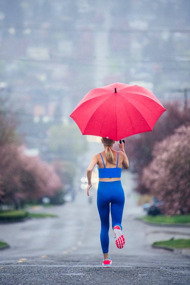 Woman running outdoors on a rainy urban street with a red umbrella and pink blossoms, USA.