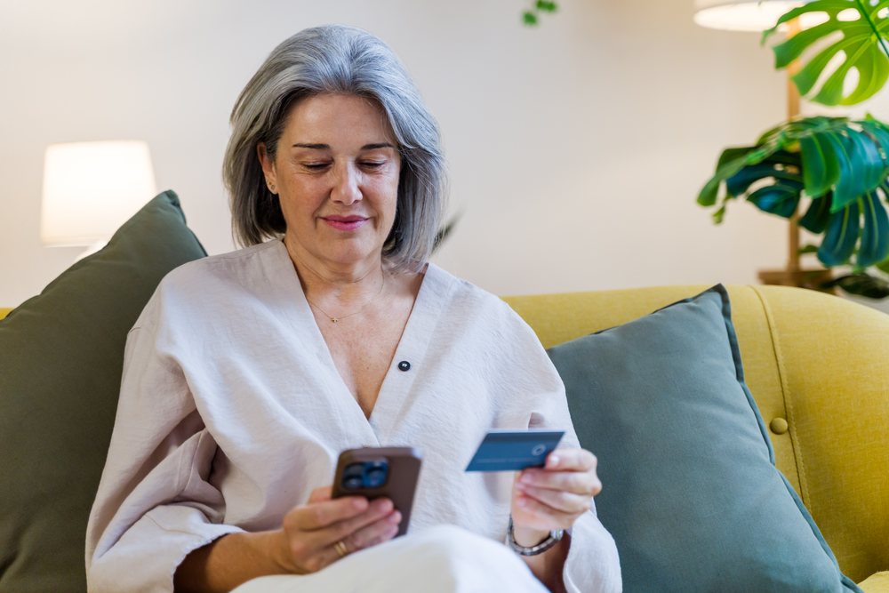 Woman sitting on a sofa holding a smartphone and credit card indoors in a modern living room with a lamp and houseplant, Spain.