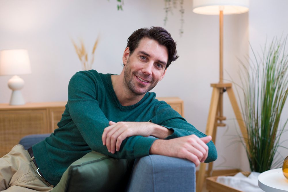 Smiling young man in green sweater sitting on a gray sofa in a modern living room, technology, young adult, home.