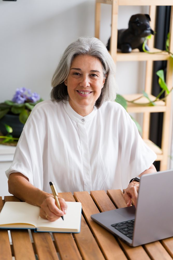 Middle aged woman writing in a notebook while using a laptop at a wooden table in a home office setting, Spain.