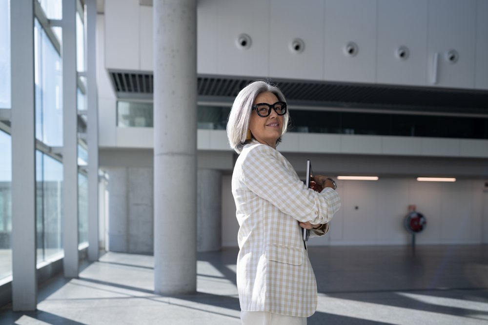 Businesswoman with gray hair and glasses holding a tablet inside a modern building with concrete pillars and natural light, Spain, Madrid.