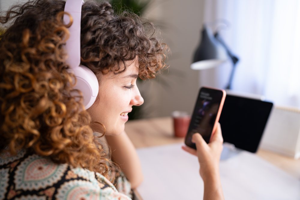 Girl with curly hair and large over-ear headphones looks at her smartphone while sitting at a desk with a tablet and desk lamp in a home office, Spain.