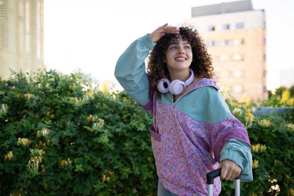 Young woman with curly hair smiling outdoors in an urban setting while holding a suitcase handle and shading her eyes from the sun, Spain.