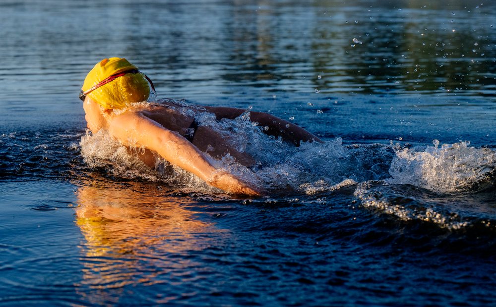 Swimmer wearing a yellow swim cap glides through open water creating splashes and reflections in the water surface, USA.